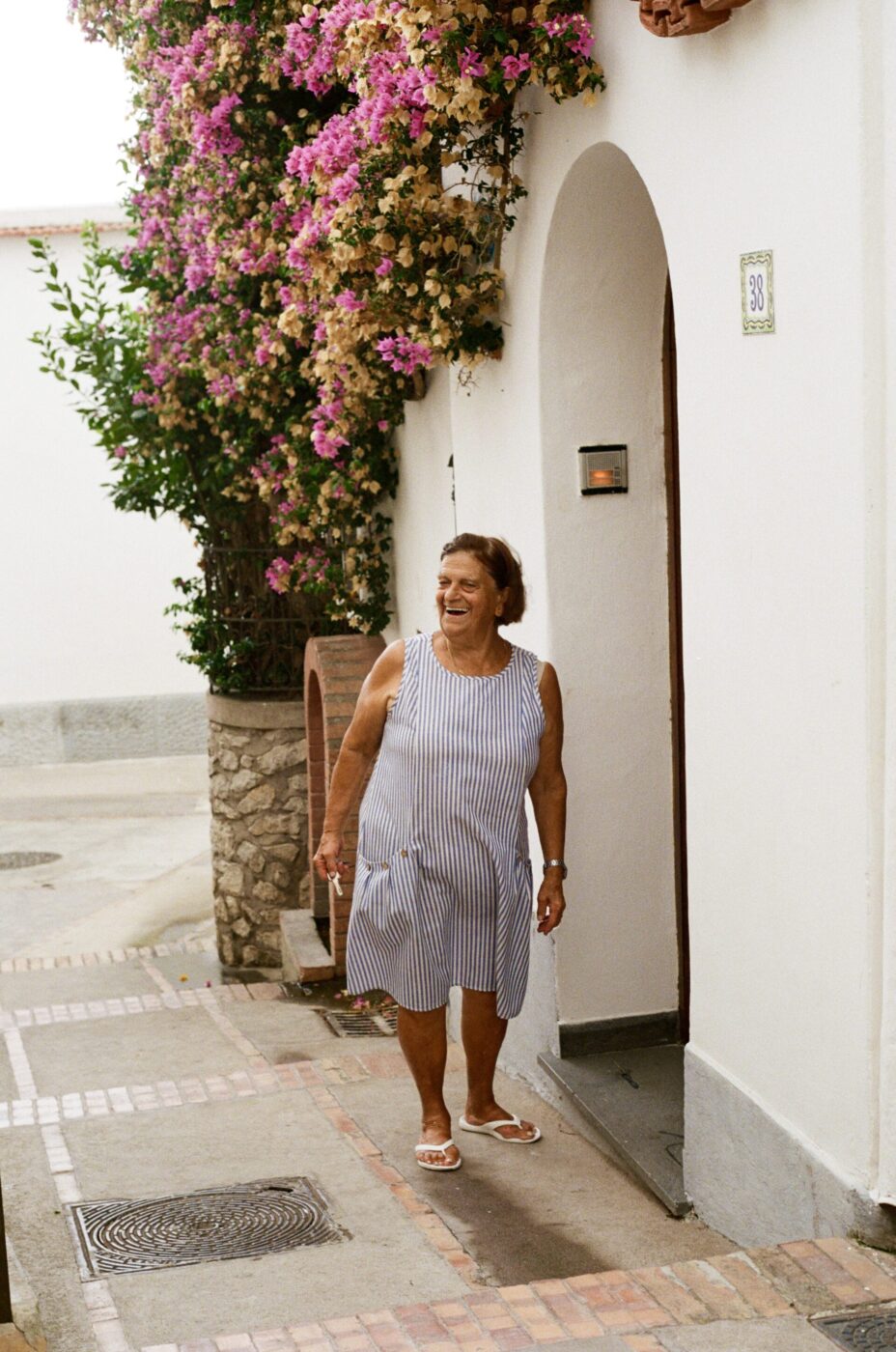 Una donna anziana in abito a righe e sandali sorride fuori da un edificio bianco con una porta ad arco, vicino a fiori colorati in fiore.