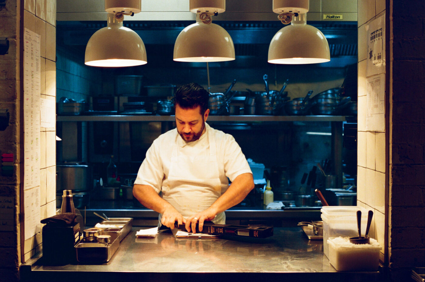 A chef in white prepares food at a steel counter under three large lamps, pots and pans on shelves visible behind him in the kitchen.