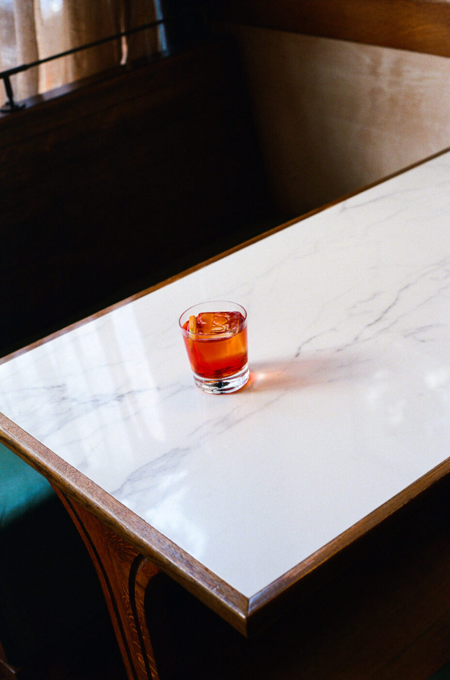 A red cocktail with ice on a white marble table in a wooden booth, sunlight casting across from the side.