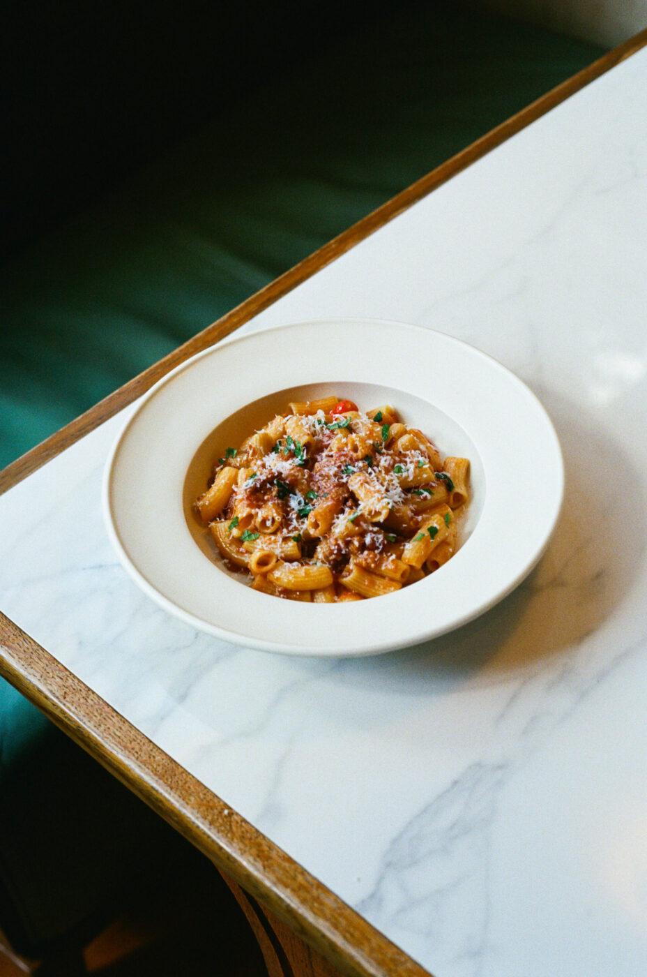 A white bowl of rigatoni in red sauce with grated cheese and herbs sits on marble beside a green-cushioned bench.