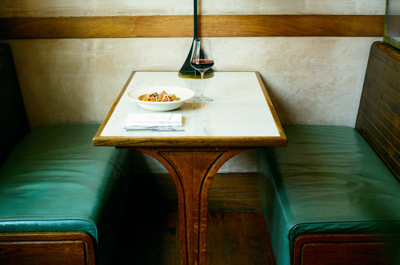 A marble-top table with green leather booth seats, set with food, red wine, and a napkin in a cozy wood-accented restaurant.