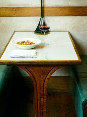 A marble-top table with green leather booth seats, set with food, red wine, and a napkin in a cozy wood-accented restaurant.