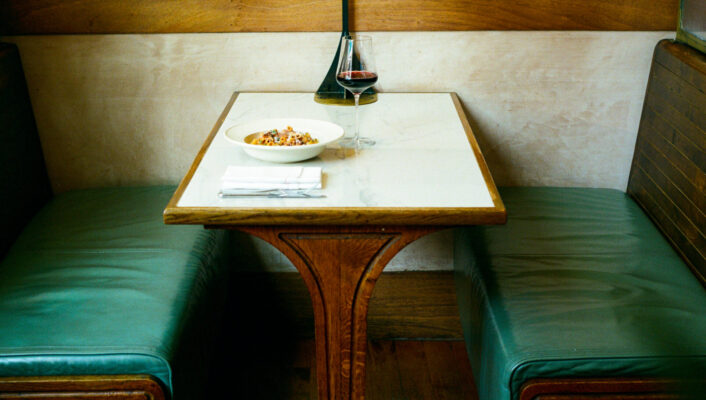 A marble-top table with green leather booth seats, set with food, red wine, and a napkin in a cozy wood-accented restaurant.