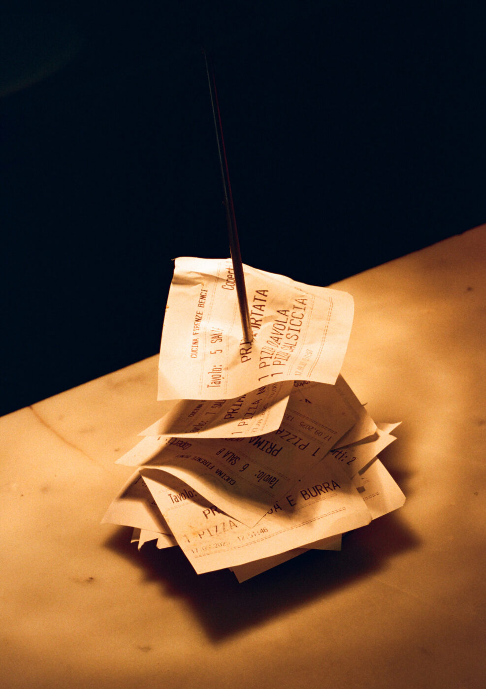 A stack of crumpled receipts skewered on a metal spindle sits on marble, bathed in warm light.