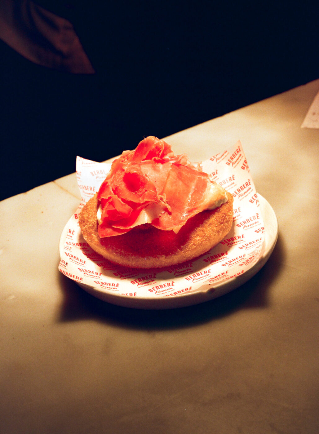 A round piece of fried bread with thin cured ham slices rests on branded paper, served on a white plate on a pale table.