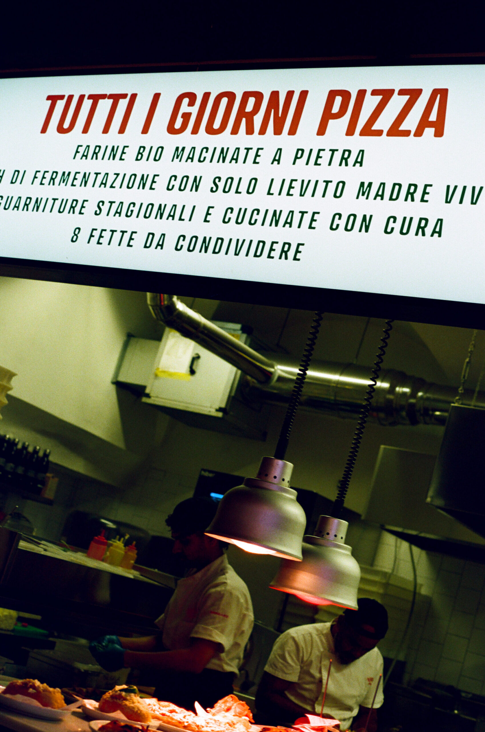 A restaurant kitchen with chefs under hanging lights; above, a sign in Italian advertises pizza with organic, natural ingredients.