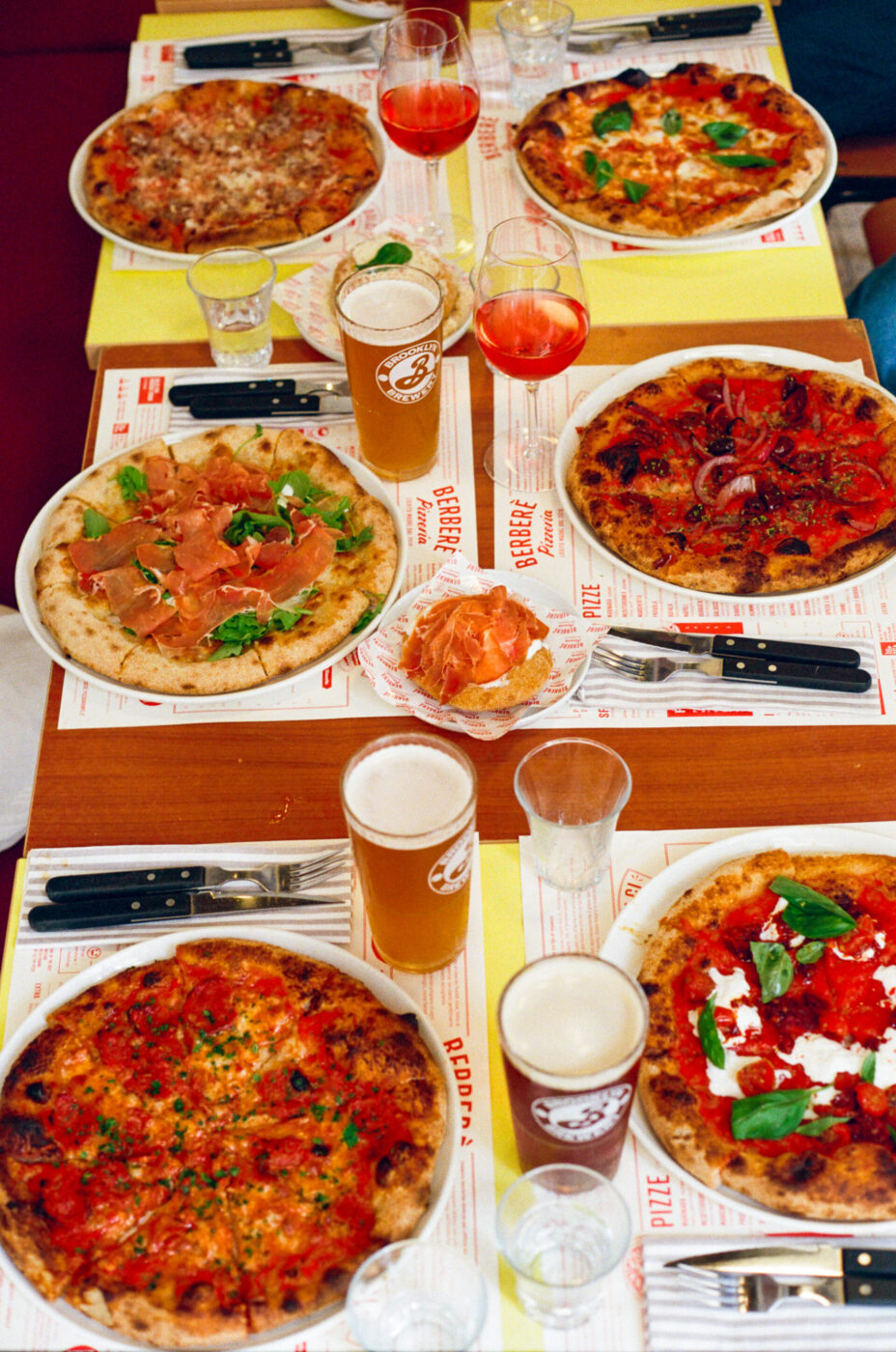 A table with six assorted pizzas, glasses of beer, red drinks, water, and chips in bowls on placemats, surrounded by cutlery.