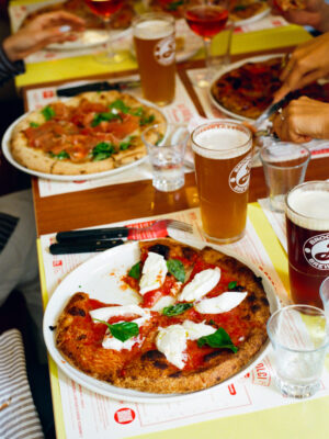 A table set with pizzas, drinks, and people reaching for slices, creating a bustling dining scene with mozzarella and basil.