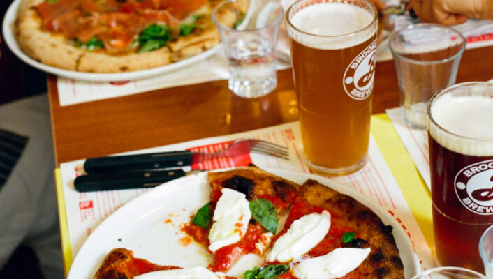 Un tavolo vivace con pizze, birra, vino e acqua, mentre le persone si avvicinano alle fette, creando un'atmosfera informale e sociale.