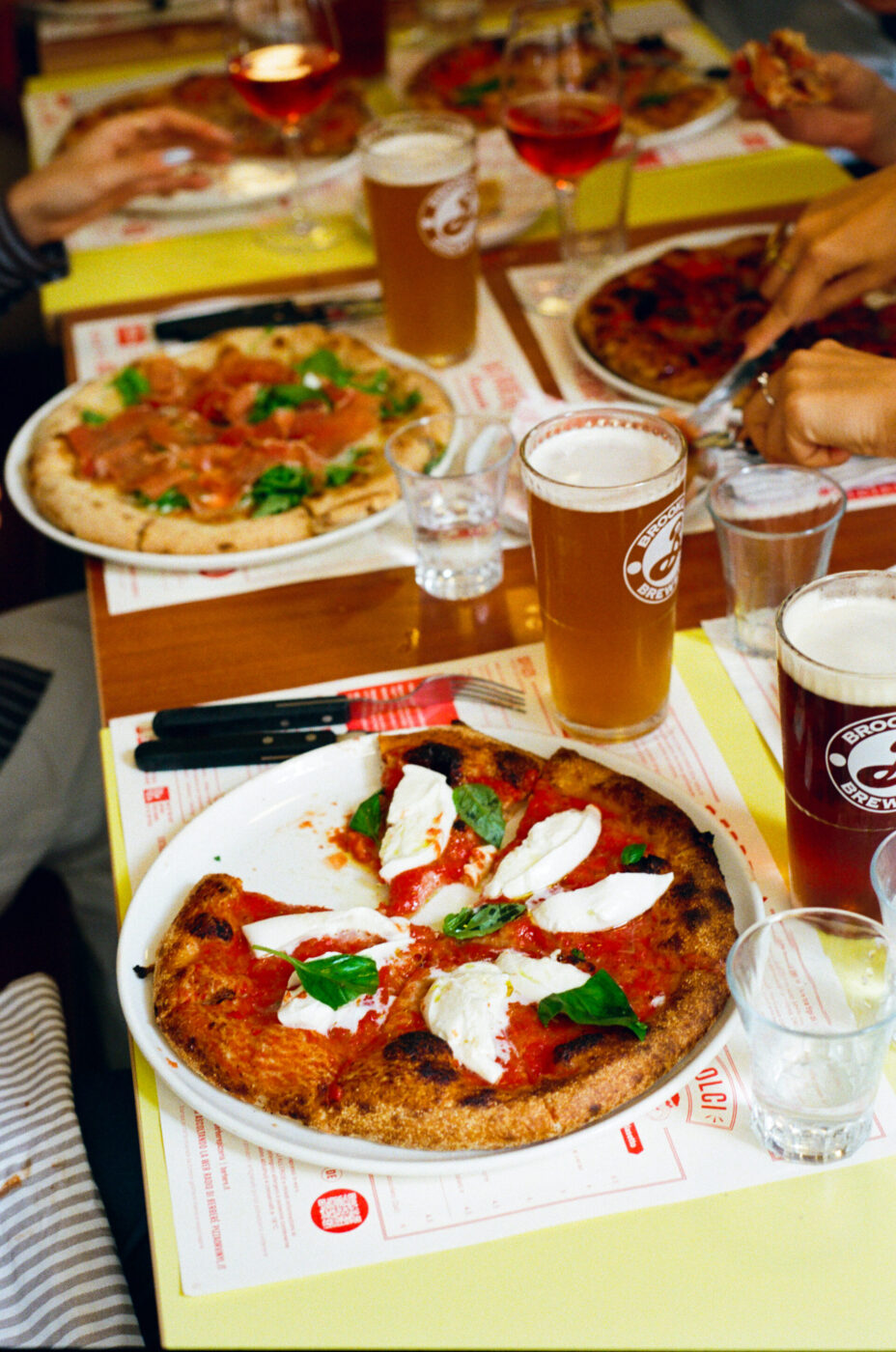 A table set with pizzas, drinks, and people reaching for slices, creating a bustling dining scene with mozzarella and basil.