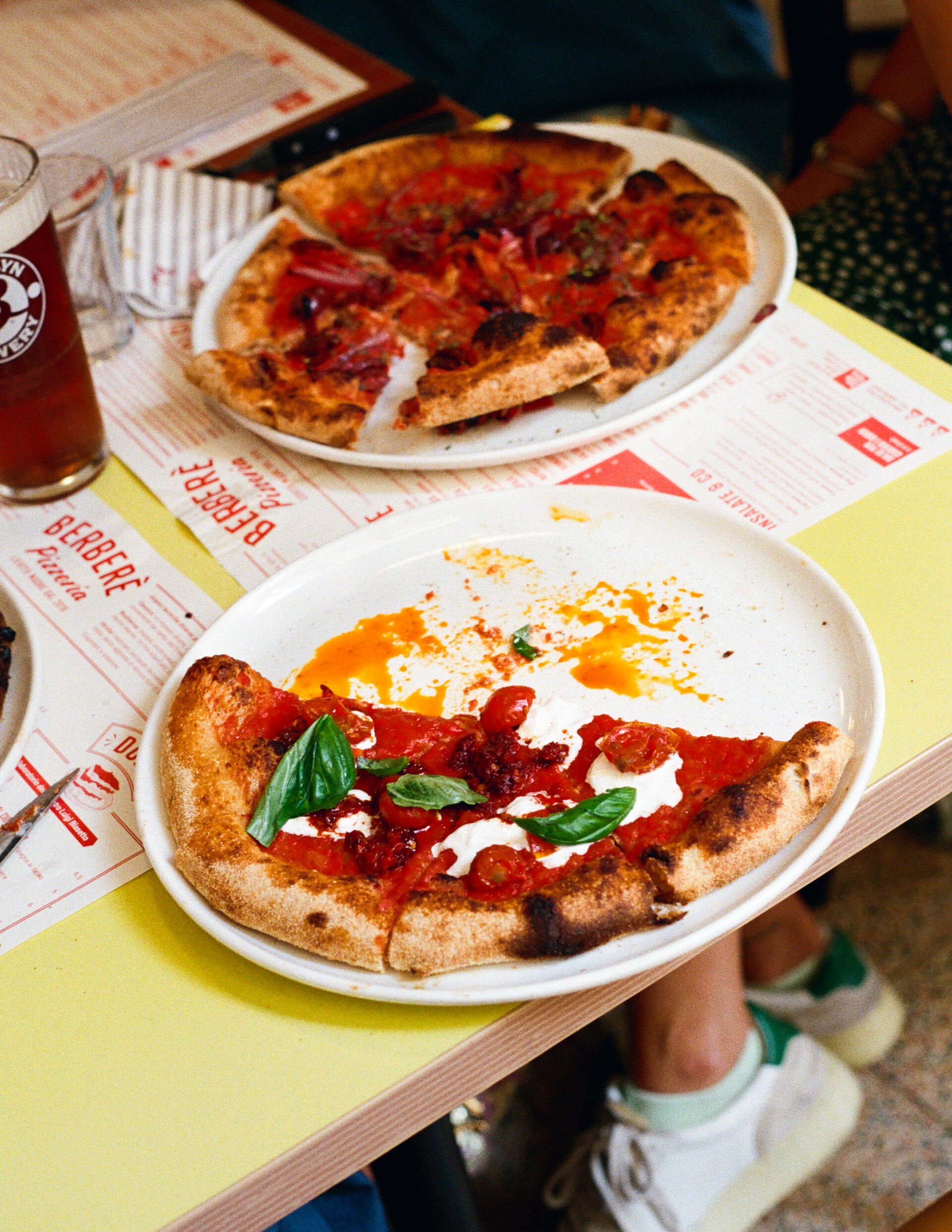 Two plates of pizza on a table, one with three slices left. Also visible: glass of beer, menus, and legs in white sneakers.