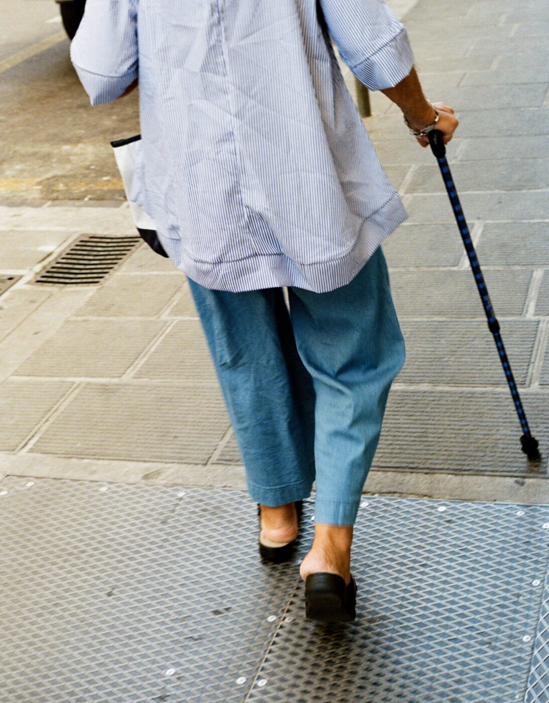 A person in a blue shirt and loose blue pants walks with a cane on a city sidewalk, seen from behind at street level.