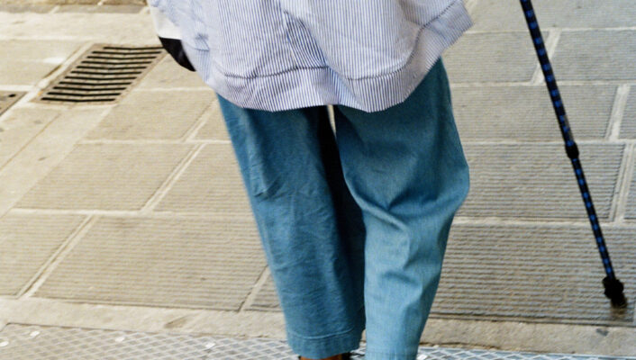 A person in a blue shirt and loose blue pants walks with a cane on a city sidewalk, seen from behind at street level.