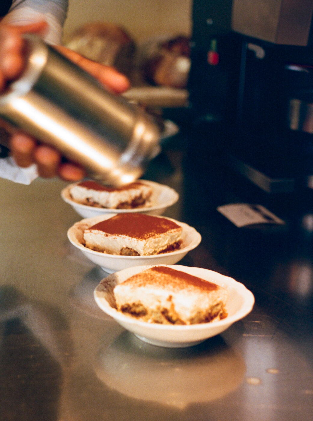A hand sprinkles cocoa from a metal shaker over three tiramisus in white bowls on a shiny counter, cream and cocoa visible on top.