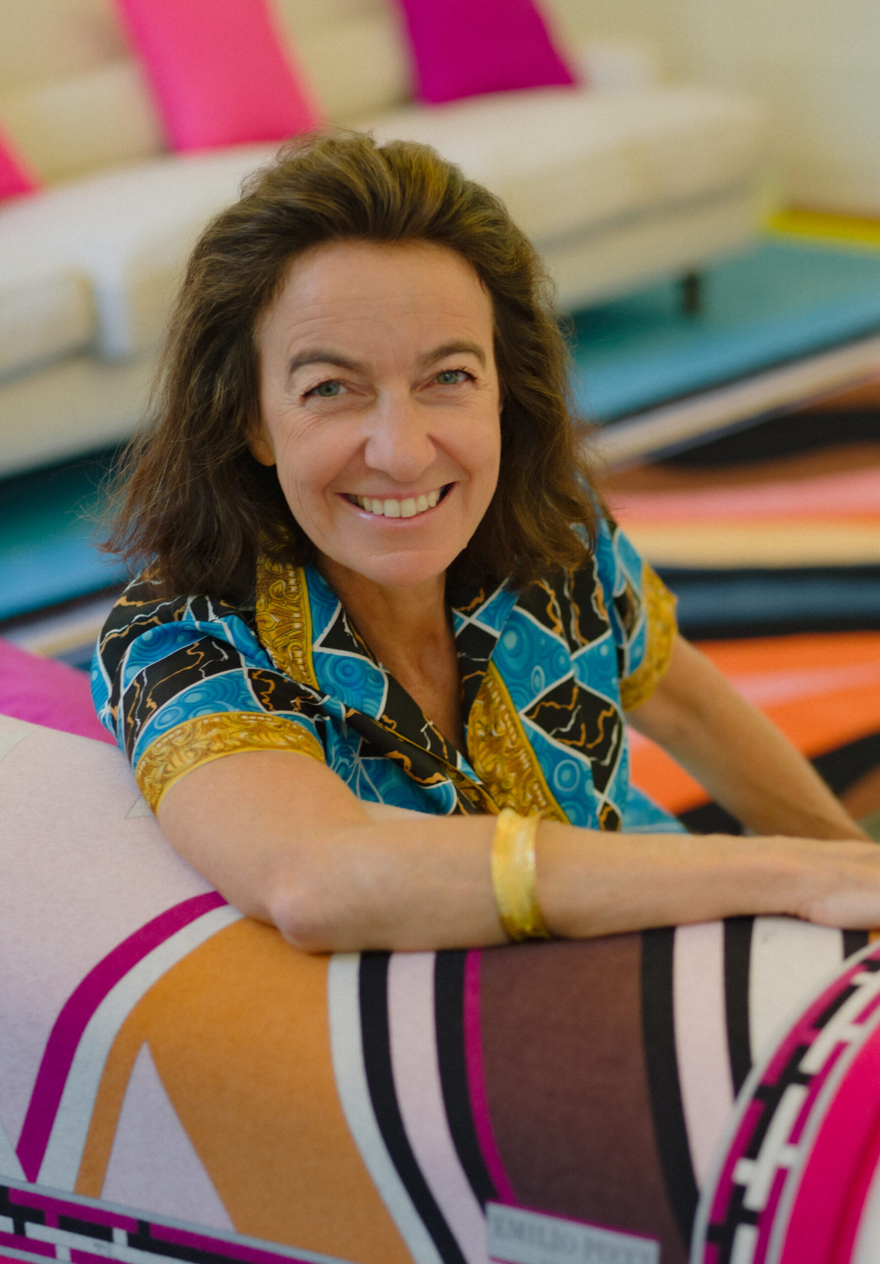 A woman with brown hair in a colorful shirt smiles on a vibrant, geometric couch; bright fabrics cover the background furniture.