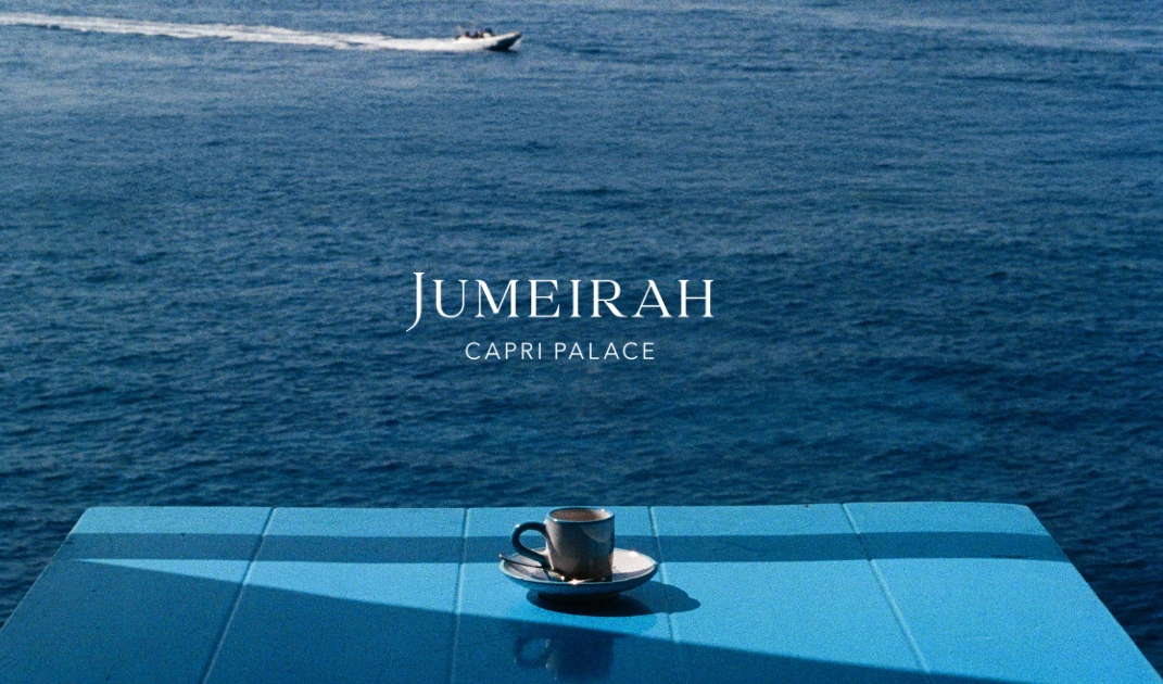 Jumeirah Capri Palace - Una tazza di caffè su un tavolo blu si affaccia sul mare, un motoscafo in lontananza, il testo del Jumeirah Capri Palace centrato.