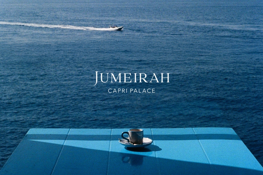 Jumeirah Capri Palace - Una tazza di caffè appoggiata su un tavolo blu acceso in riva al mare, con un motoscafo in lontananza. Testo: Jumeirah Capri Palace.