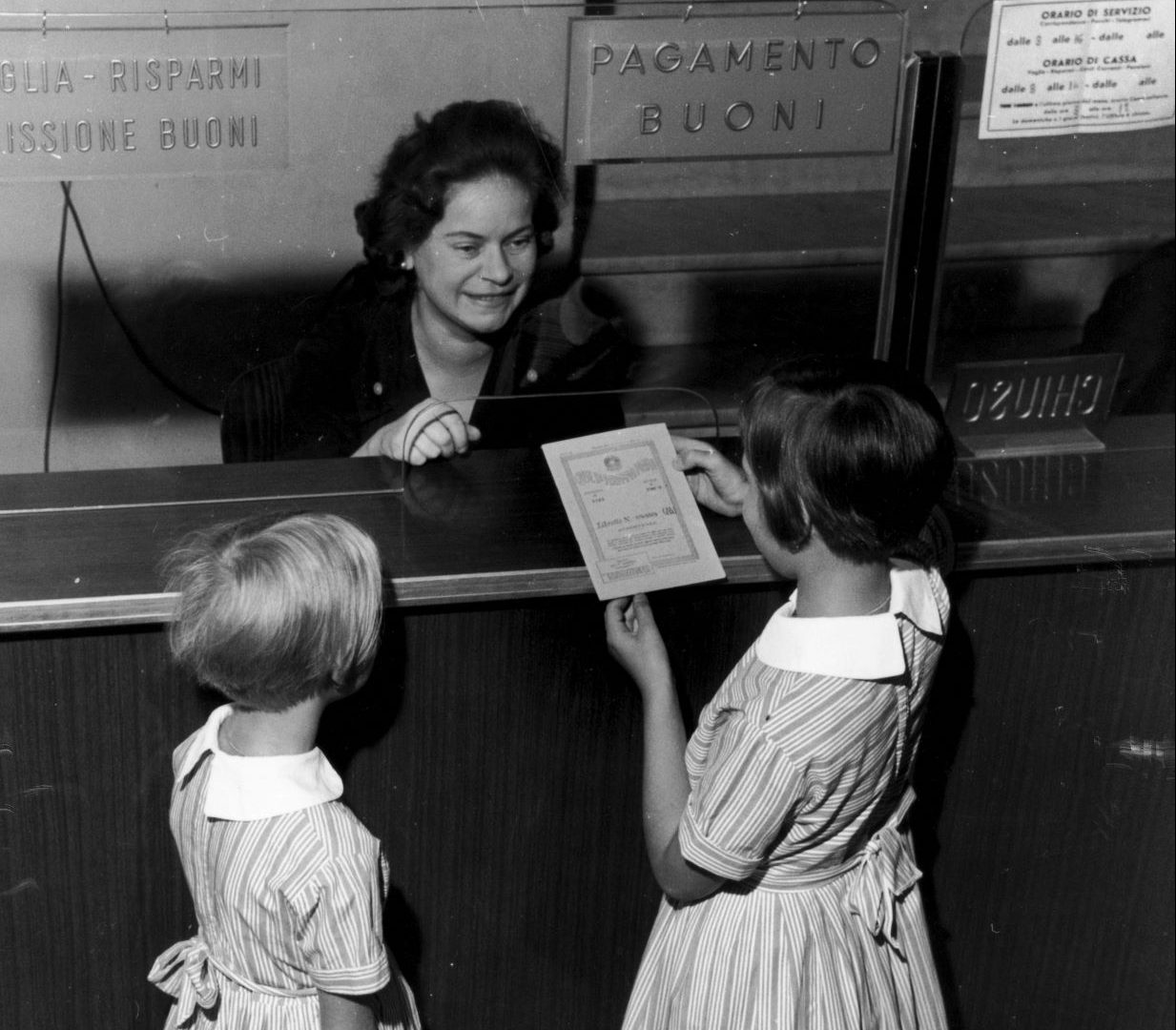 Due bambini in abiti a righe consegnano un documento a una cassiera sorridente dietro un vetro; sopra di lei sono visibili le insegne italiane.
