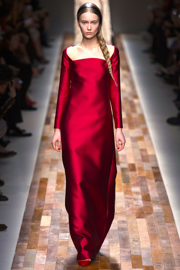A model walks a wooden runway in a long, fitted off-shoulder red satin gown. Her braid drapes forward; audience blurred behind.