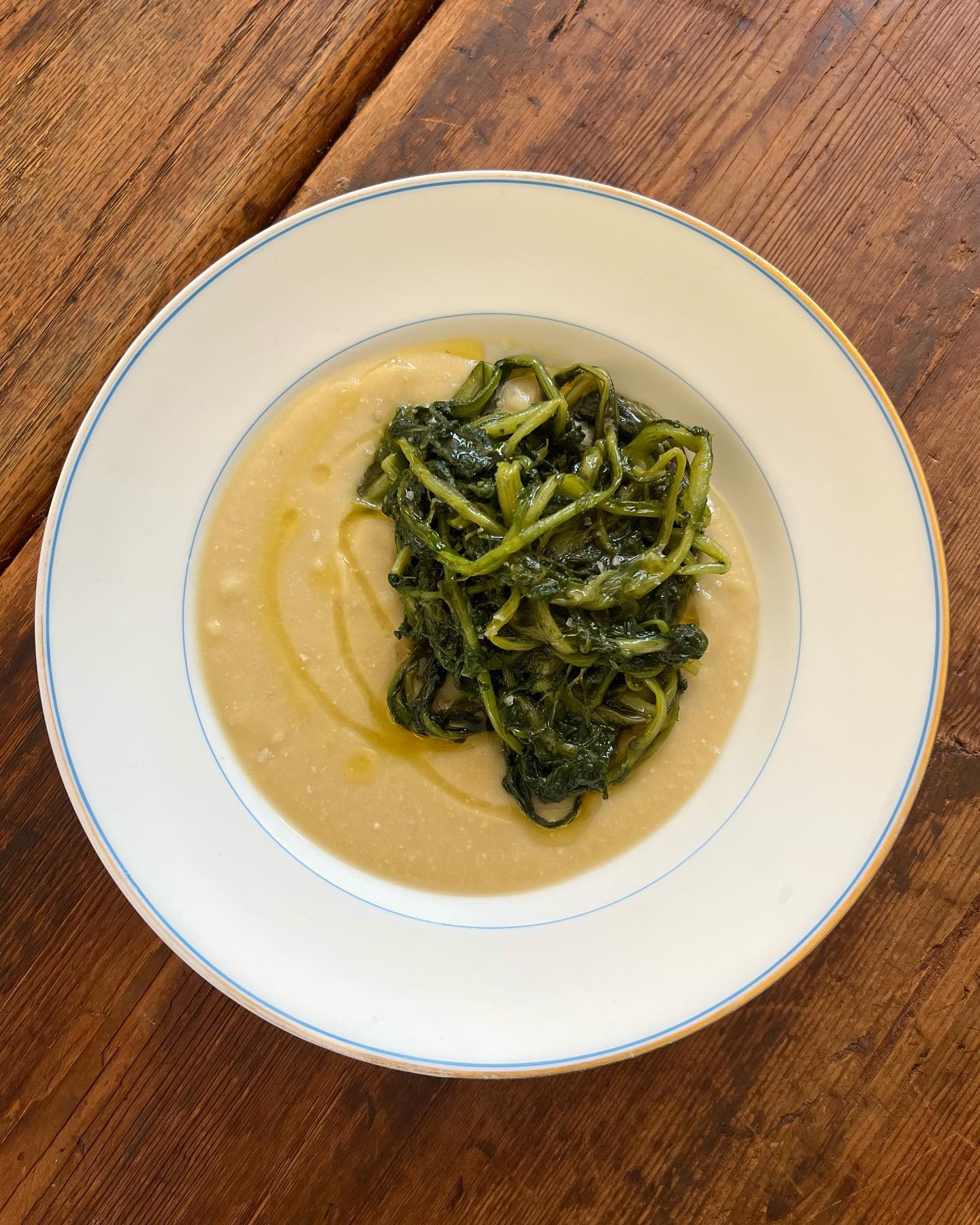 A white bowl of creamy beige purée with sautéed greens and olive oil drizzle sits on a wooden table.