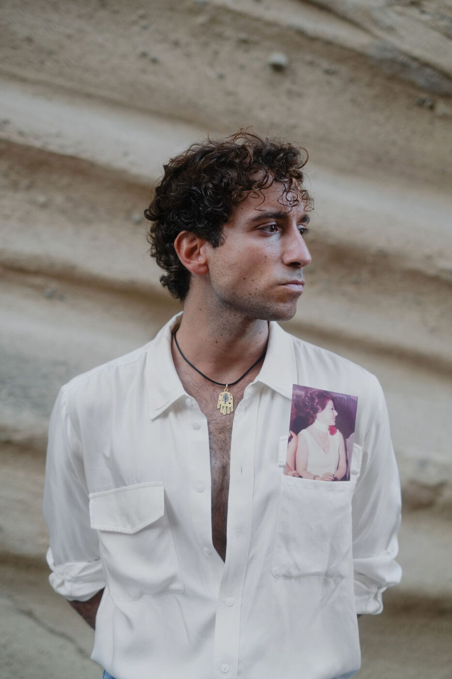 Un uomo con i capelli ricci, con una camicia bianca con una foto di donna sul taschino, è in piedi accanto a una parete beige e guarda di lato, serio.