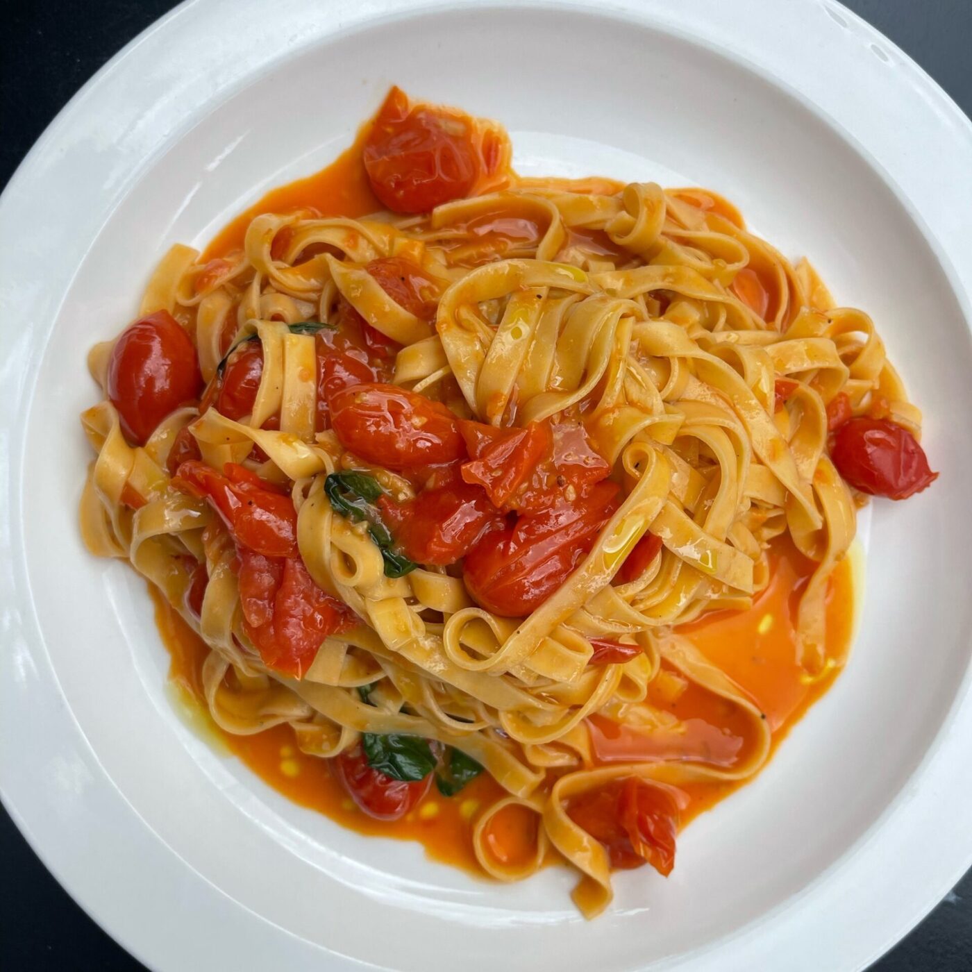 A plate of tagliatelle pasta with cherry tomatoes, basil, and light tomato sauce on a white dish. The meal looks fresh and vibrant.