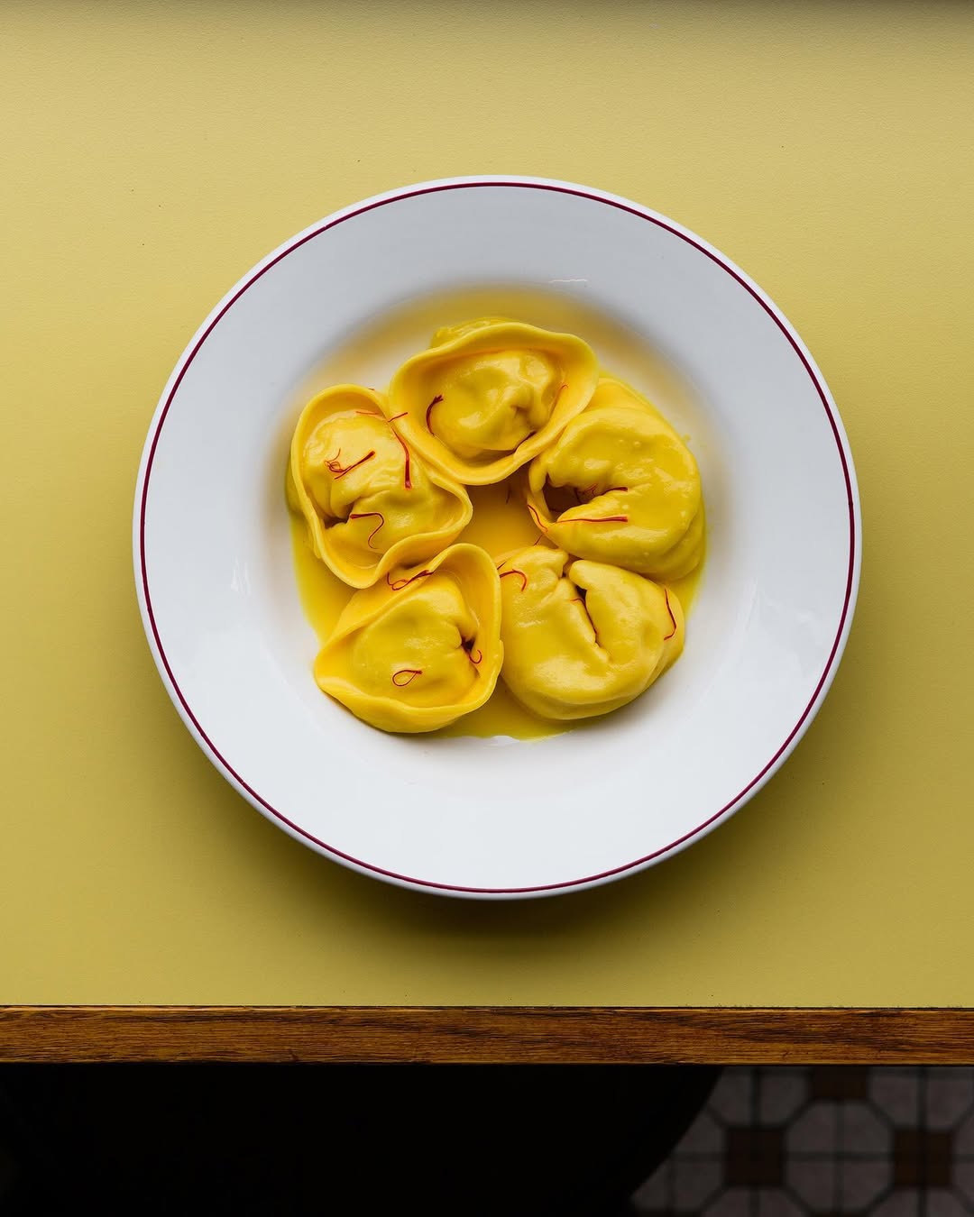 A white plate with a red rim holds five yellow tortellini in light broth, topped with saffron, resting on a yellow table.