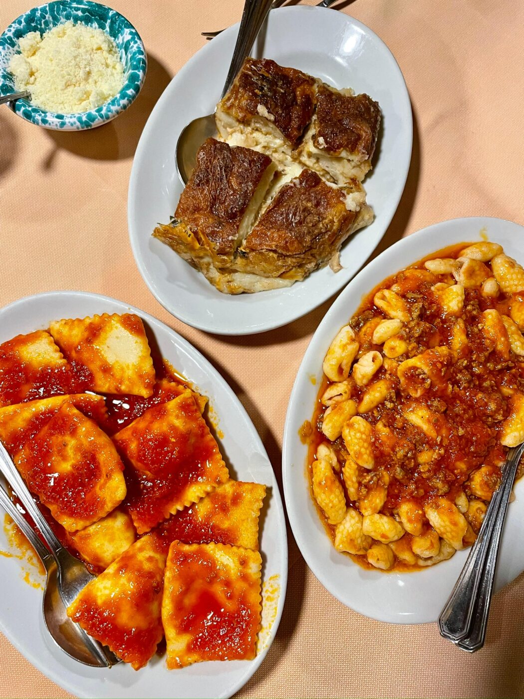 Three oval plates with ravioli, gnocchi, and lasagna on a peach cloth; grated cheese bowl and spoons beside each dish.
