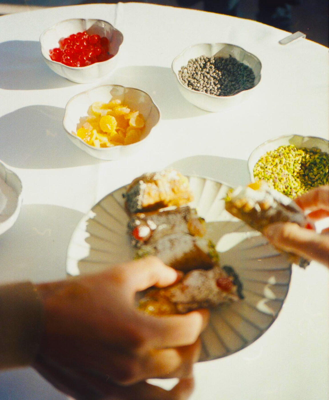 A person breaks apart a pastry over a plate, surrounded by bowls of cherries, chocolate chips, candied orange, and pistachios.