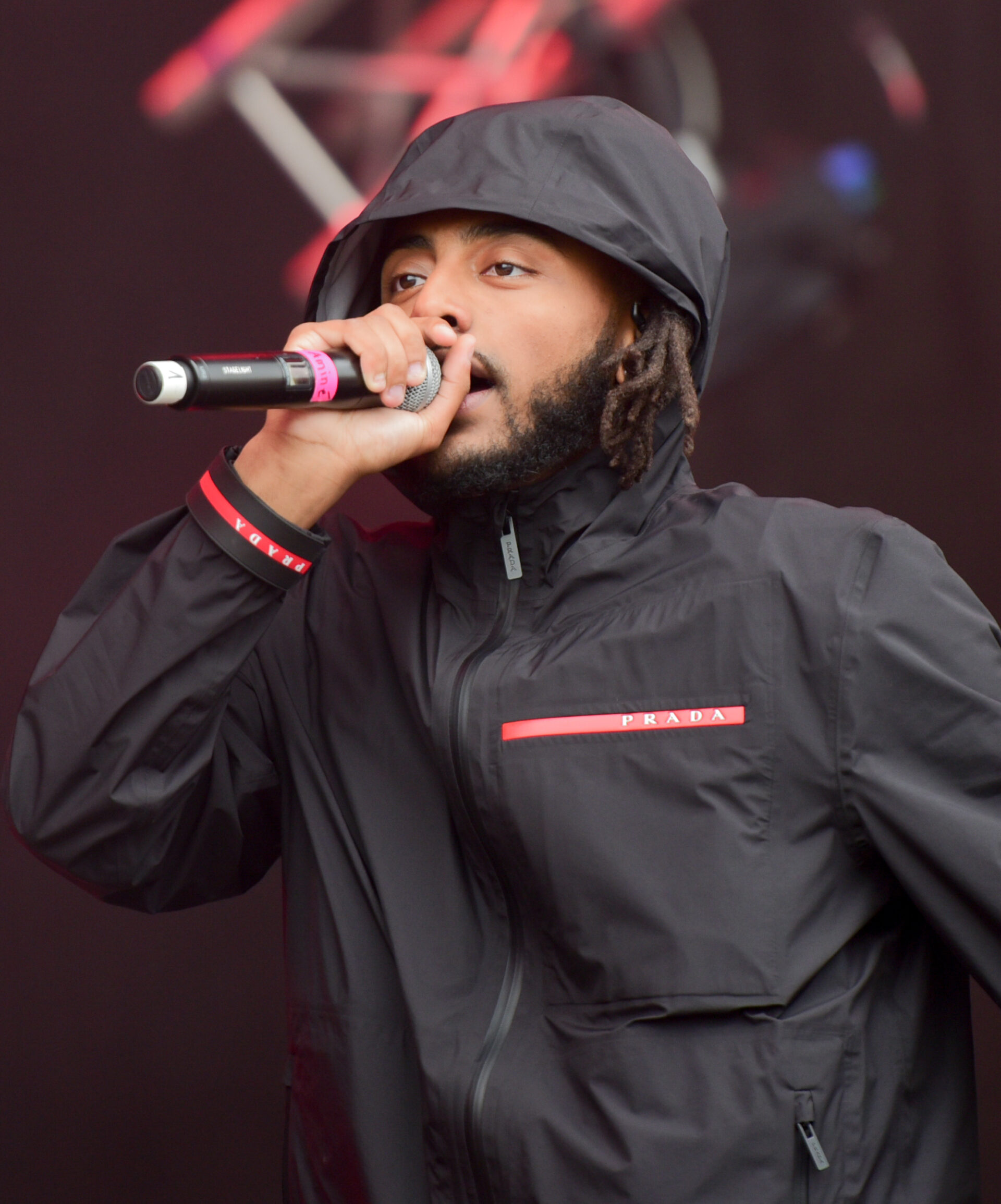 Aminé in a black Prada hooded jacket performs on stage, holding a microphone close to his mouth.