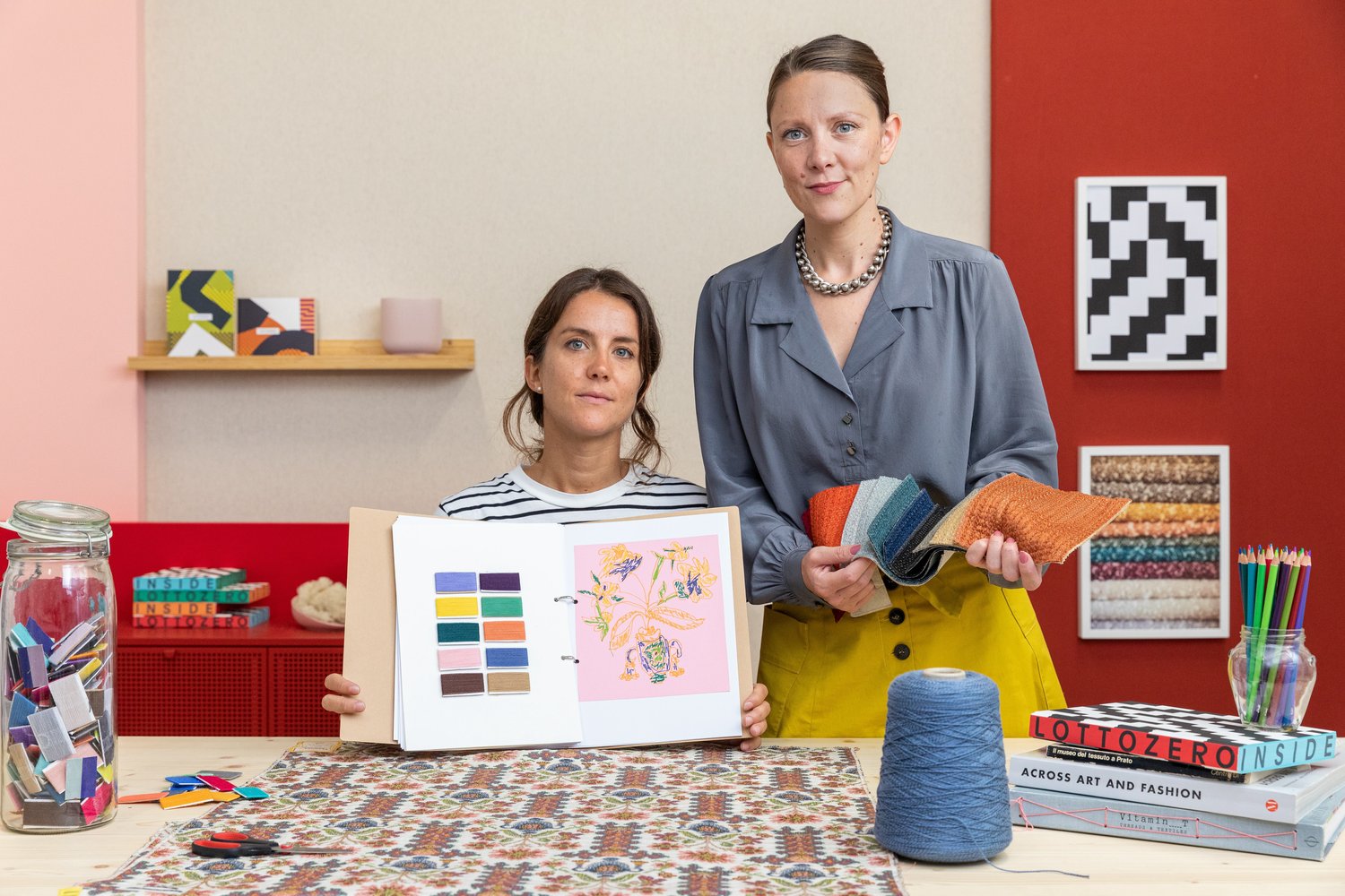 Two women in a vibrant studio display design materials; one shows swatches in a book, the other holds fabric, with supplies on the table.