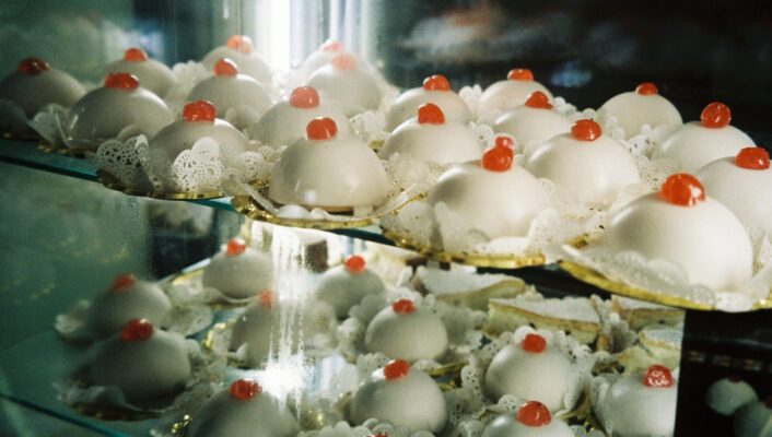 Rows of dome-shaped pastries with white icing and red cherries are lined on glass bakery shelves, each on a paper doily.