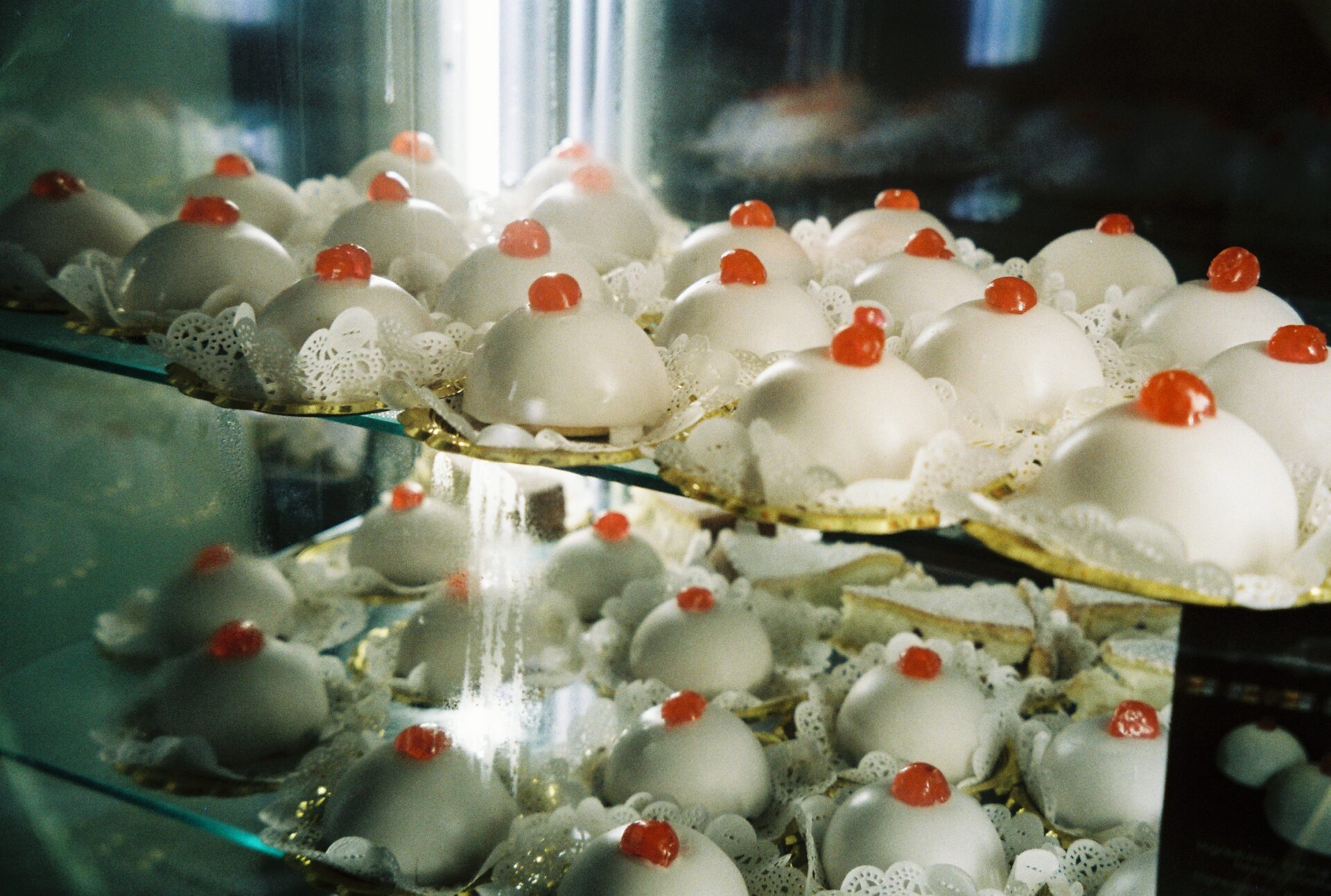 Rows of dome-shaped pastries with white icing and red cherries are lined on glass bakery shelves, each on a paper doily.