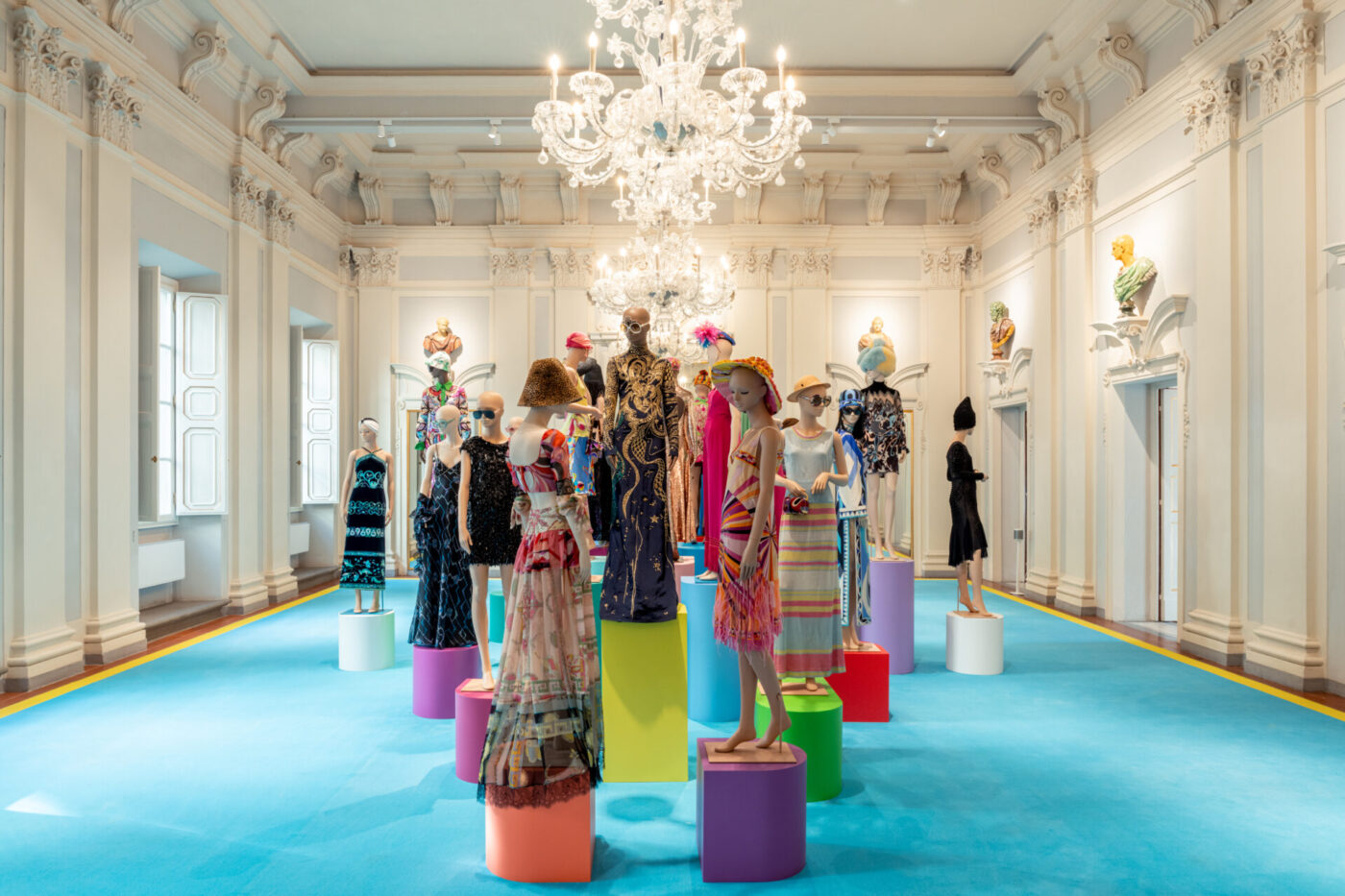 A bright, ornate gallery shows mannequins in vivid designer outfits, chandeliers above, sculptures on walls, and blue carpeted floor.