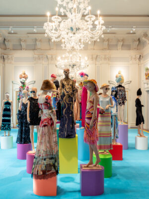 A bright, ornate gallery shows mannequins in vivid designer outfits, chandeliers above, sculptures on walls, and blue carpeted floor.