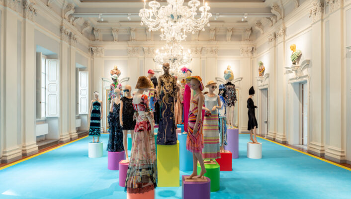 A bright, ornate gallery shows mannequins in vivid designer outfits, chandeliers above, sculptures on walls, and blue carpeted floor.