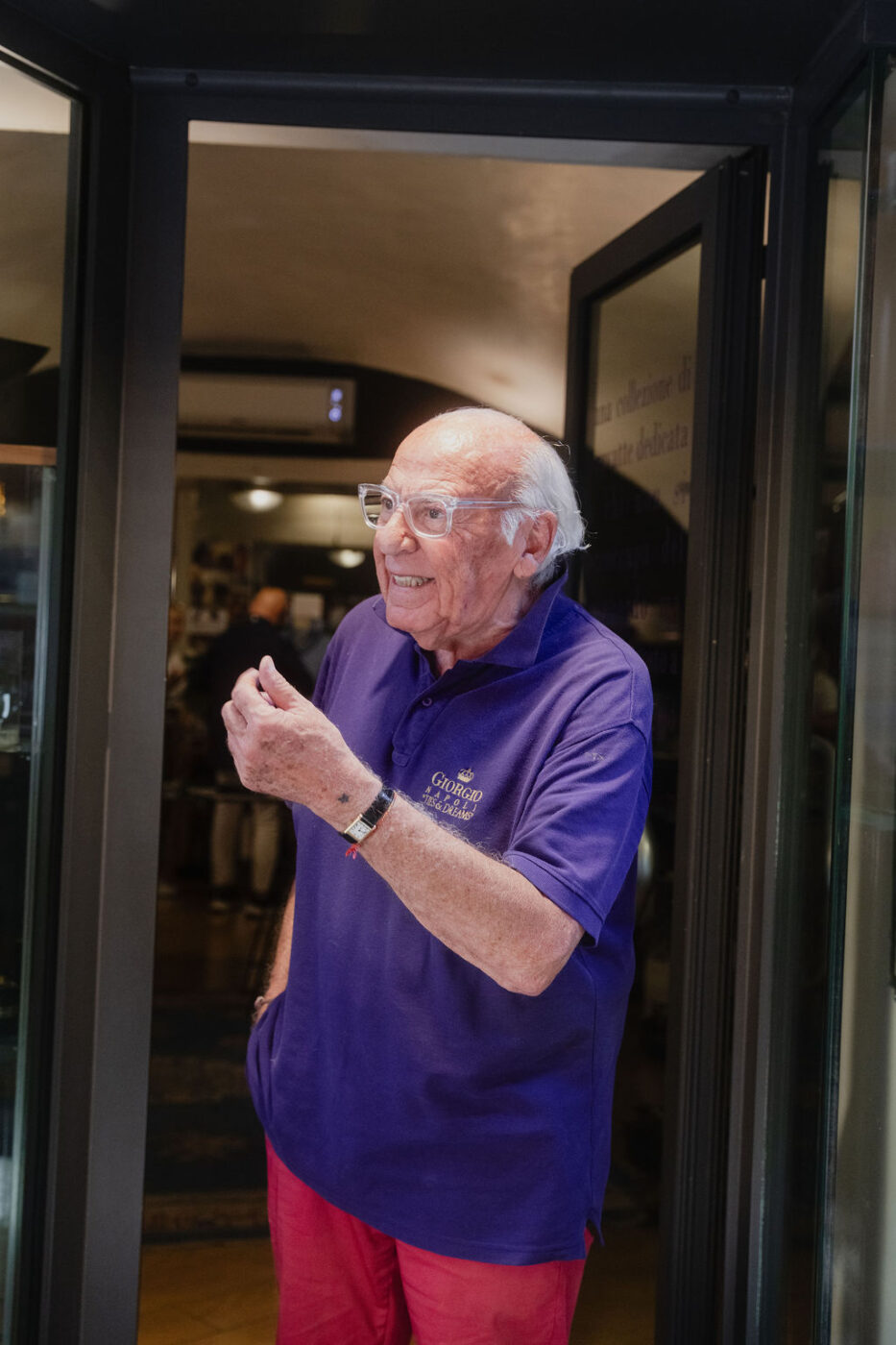 Un uomo anziano con gli occhiali, in polo viola e pantaloni rossi, sta in piedi sorridendo all'ingresso, gesticolando con una mano.