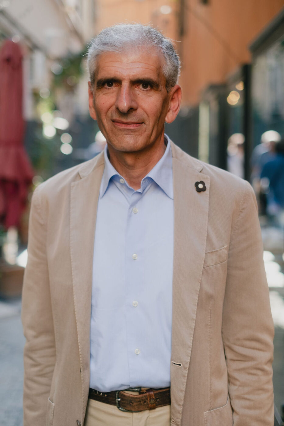 Un uomo anziano con i capelli corti e grigi, in blazer beige e camicia blu, sta all'aperto e guarda la telecamera alla luce del sole.