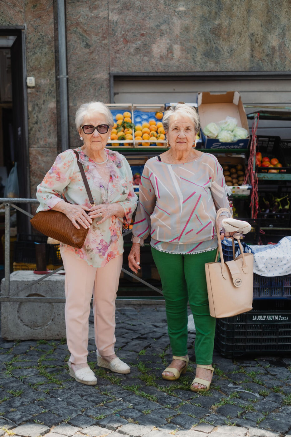 Due donne anziane, con occhiali da sole e abbigliamento casual, sono in piedi accanto a una bancarella di frutta e verdura su una strada acciottolata.