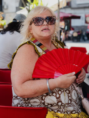 Una donna con i capelli biondi, con gli occhiali da sole e un vestito a fantasia, siede all'aperto con un ventaglio rosso in mano su una sedia rossa, in mezzo a persone e ombrelli.