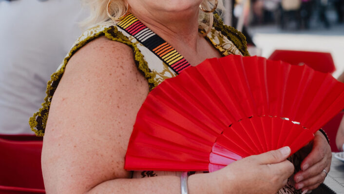 A blonde woman in large sunglasses holds a red fan outdoors, wearing a patterned dress; red chairs and a busy street are behind her.