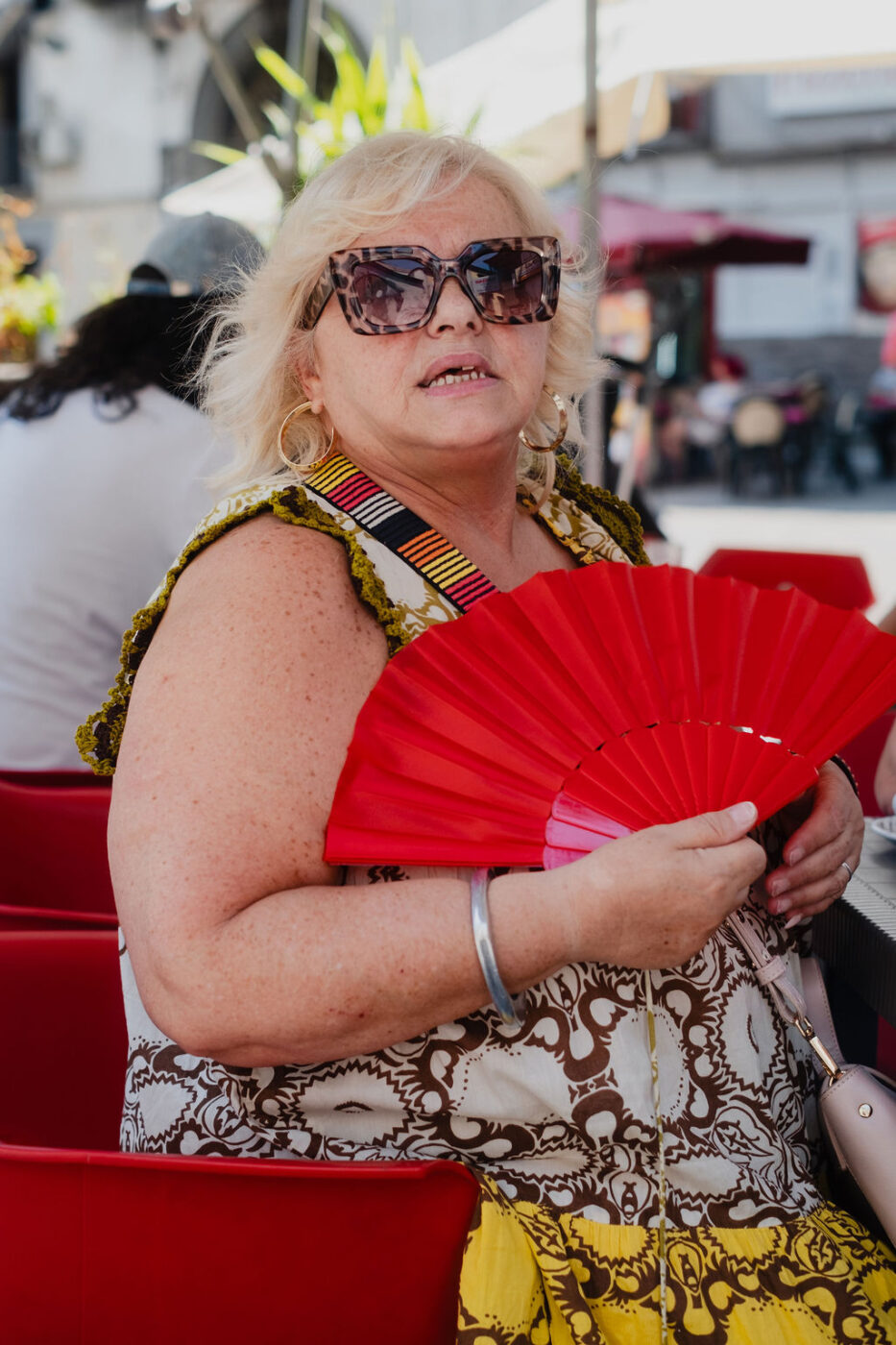 Una donna con i capelli biondi, con gli occhiali da sole e un vestito a fantasia, siede all'aperto con un ventaglio rosso in mano su una sedia rossa, in mezzo a persone e ombrelli.