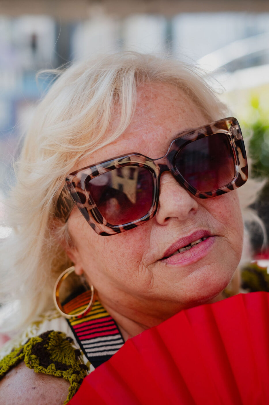 Una donna anziana con i capelli biondi, gli occhiali da sole tartarugati e gli orecchini a cerchio dorati regge un ventaglio rosso, con la luce del sole sul viso.