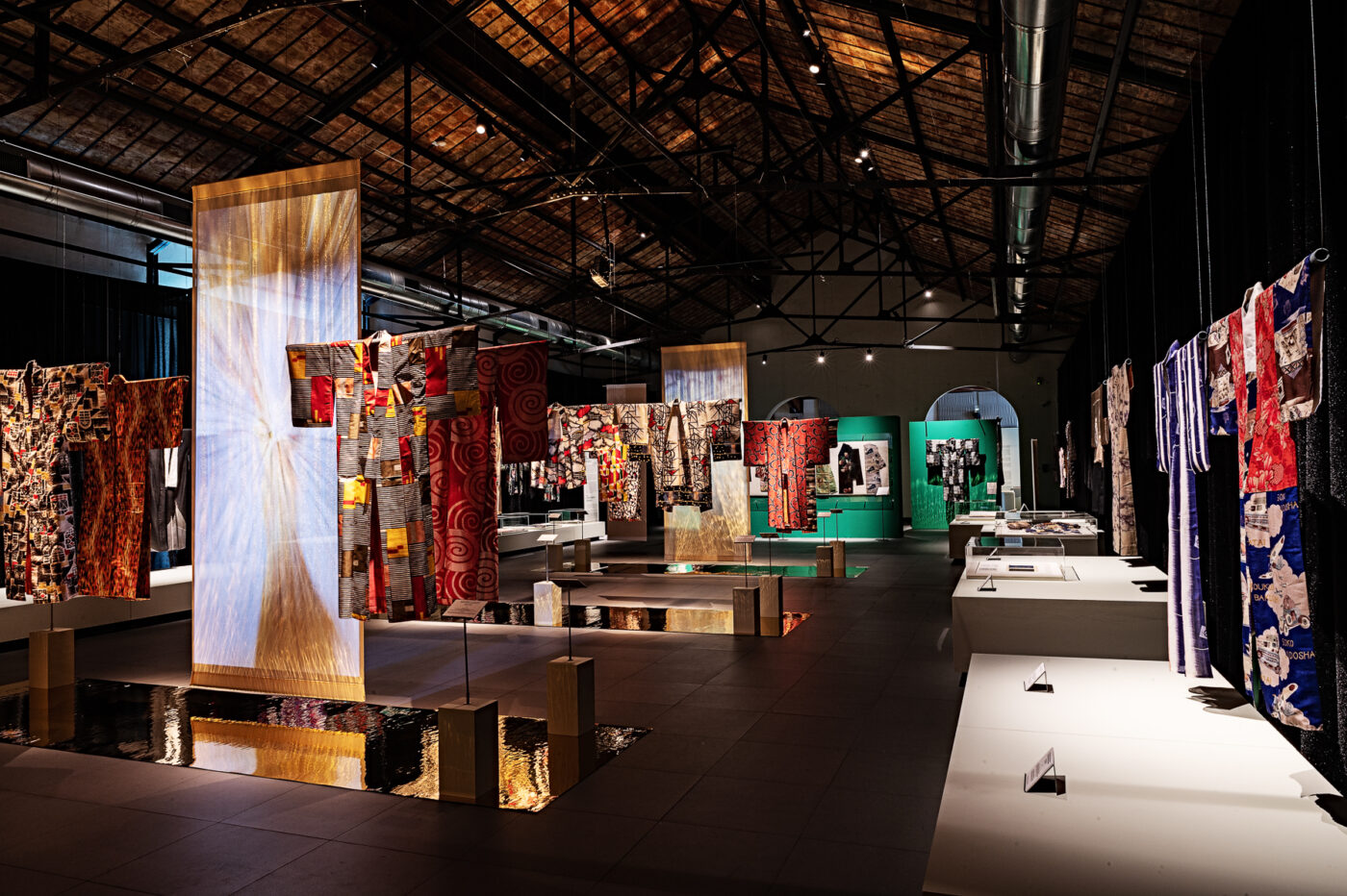 Colorful kimonos on stands in a modern hall with high, exposed-beam ceilings and dramatic lighting, creating a vibrant atmosphere.