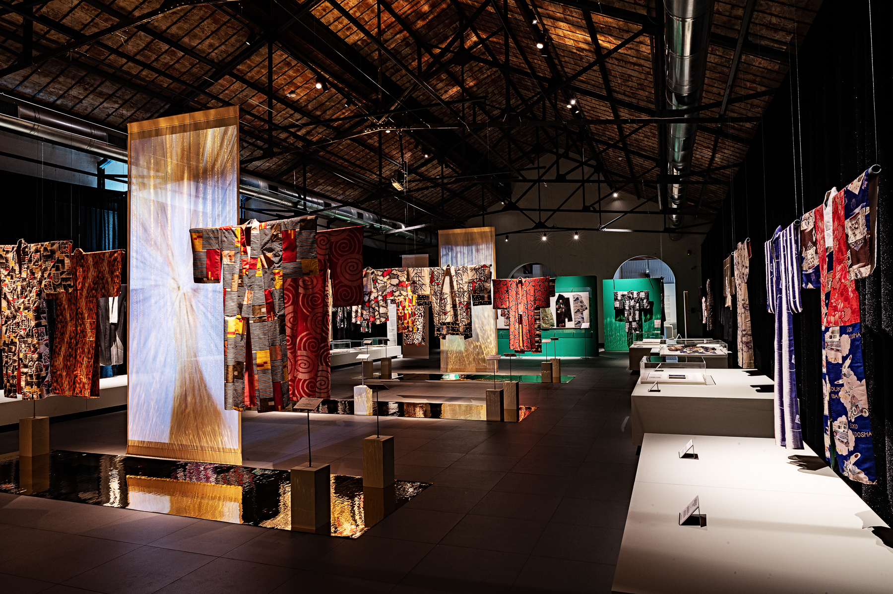 Colorful kimonos on stands in a modern hall with high, exposed-beam ceilings and dramatic lighting, creating a vibrant atmosphere.