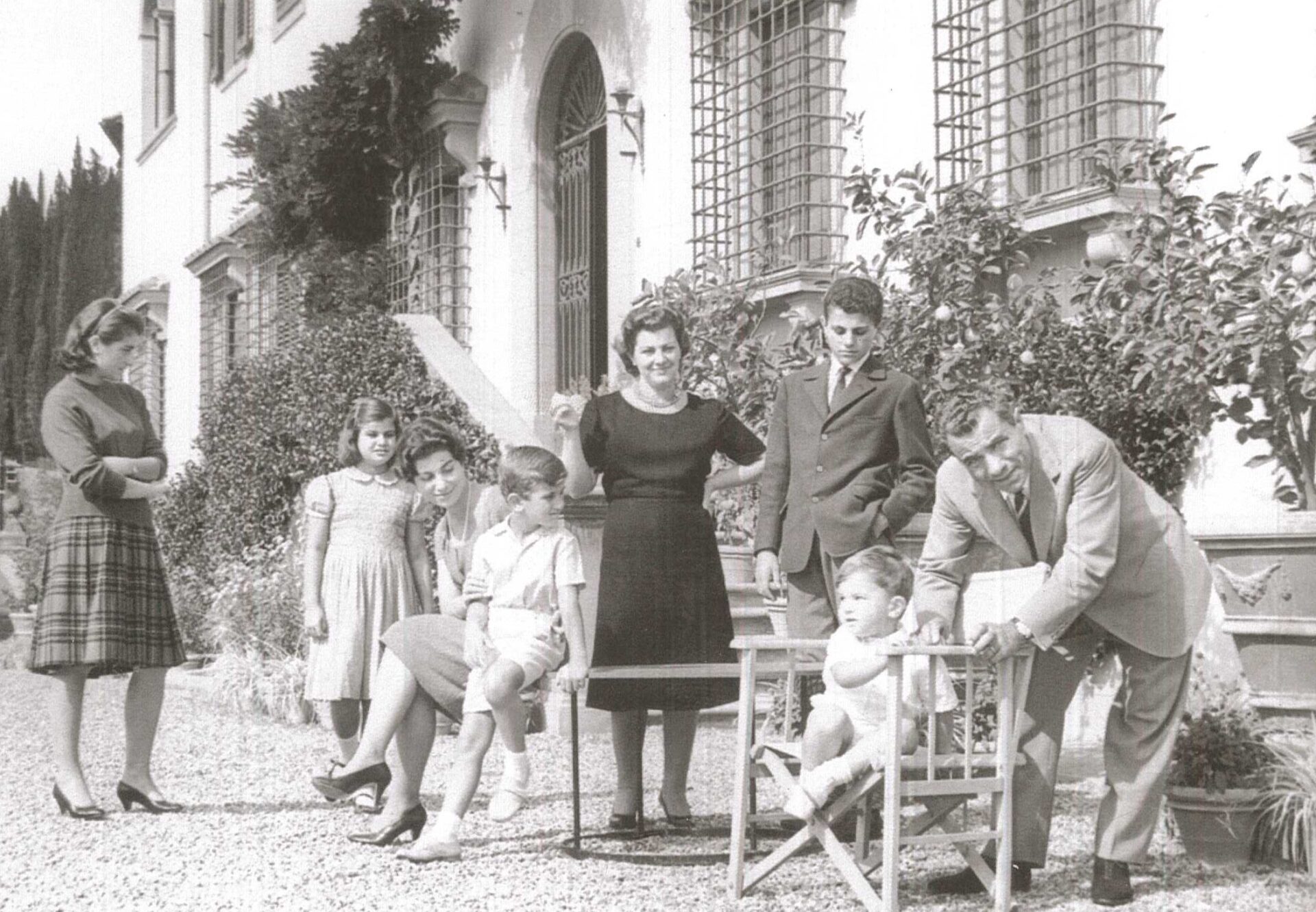A black-and-white photo of a family outside a large building, with adults and kids by lush plants and stairs.