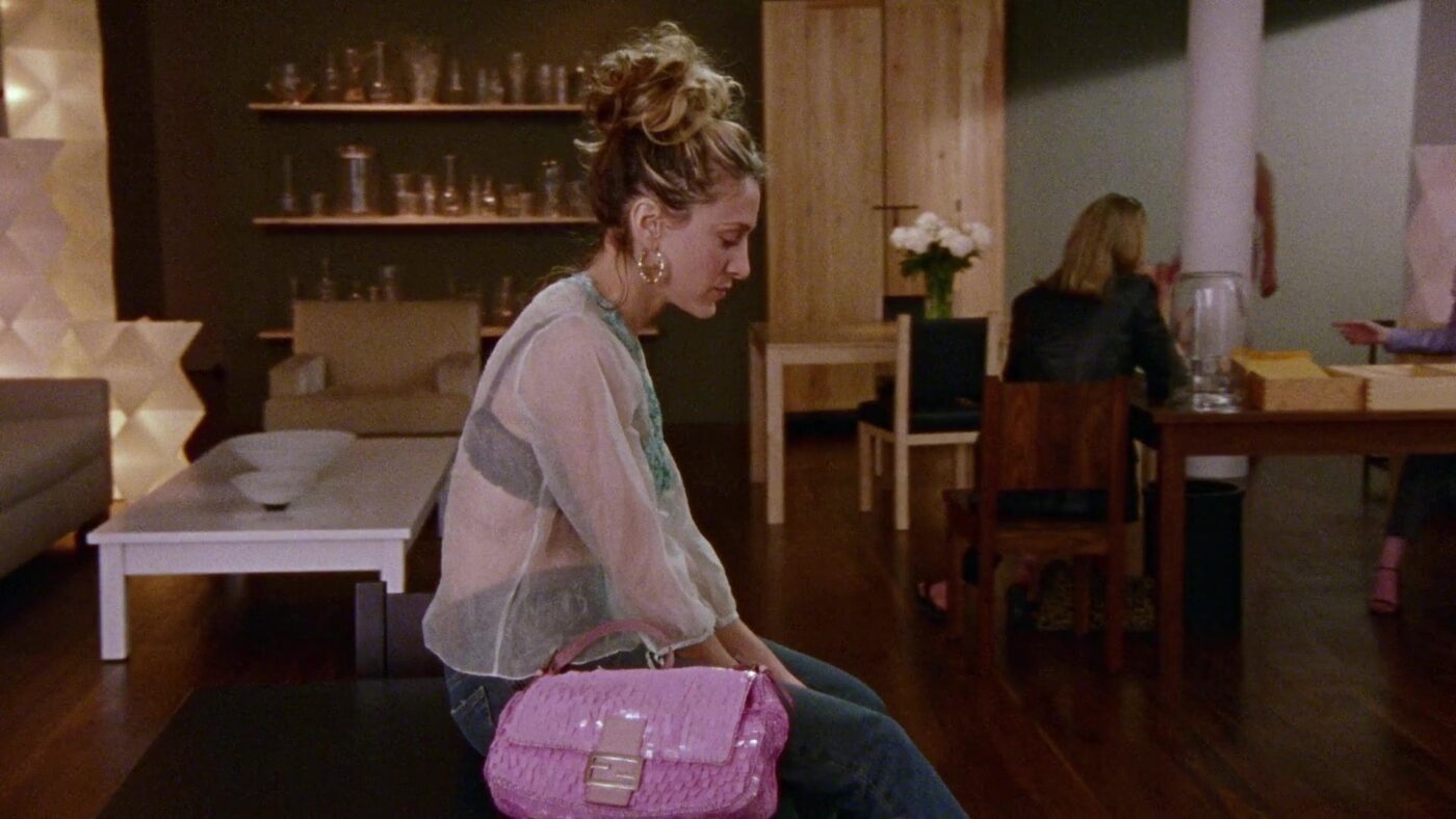 a still from Sex and the City; Carrie with curly hair in a messy bun sits on an indoor bench, looking down, wearing a sheer white top, jeans, and holding a pink handbag.