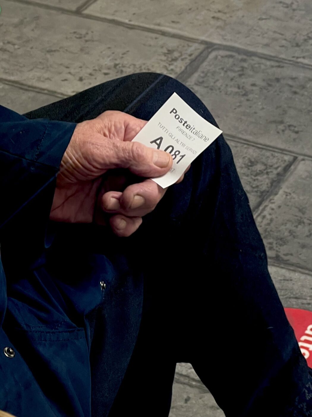 Una persona tiene in mano il biglietto di coda A081 di Poste Italiane, con la mano appoggiata sul ginocchio, sopra una pavimentazione in piastrelle grigie.