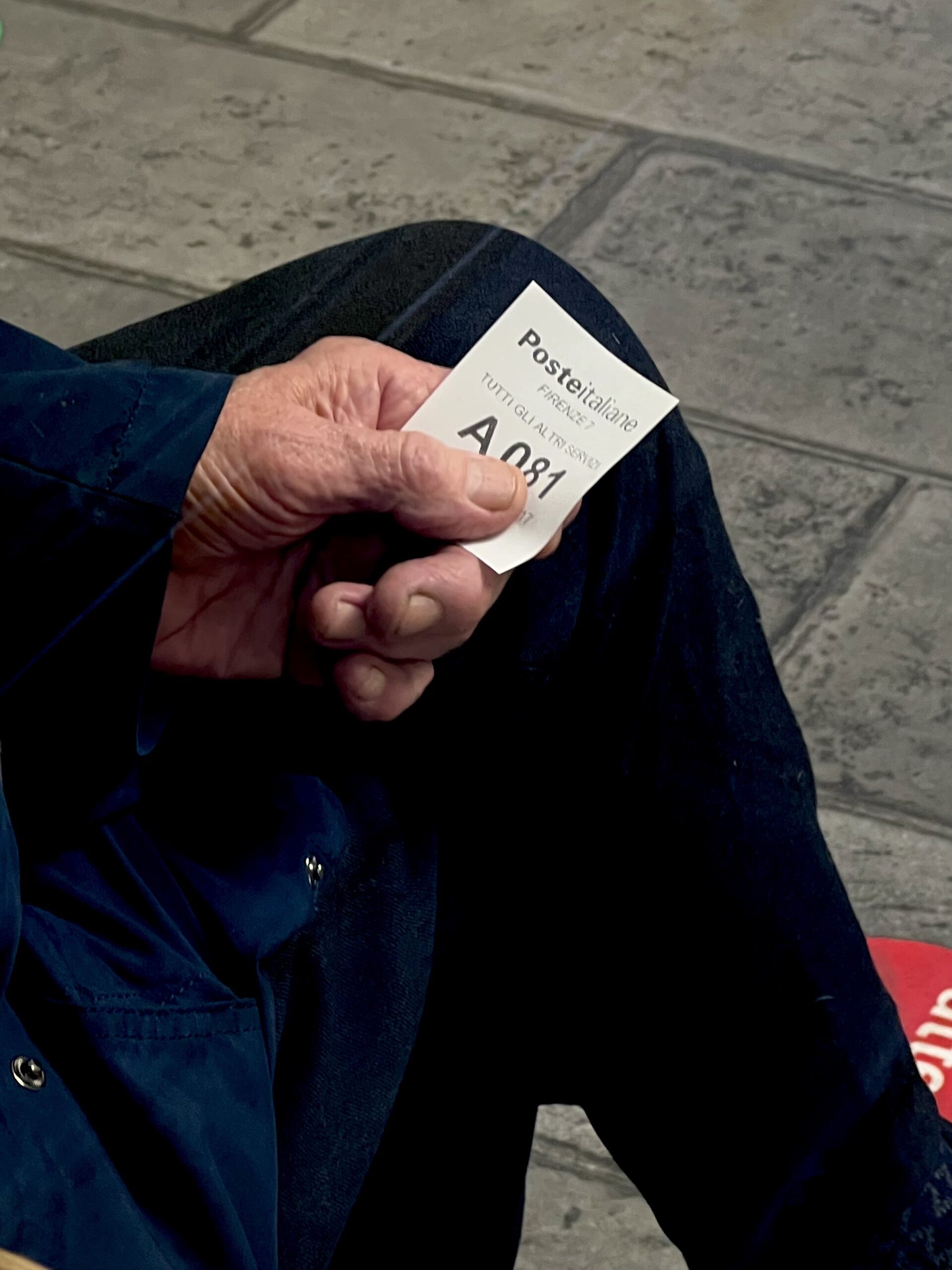 Una persona tiene in mano il biglietto di coda A081 di Poste Italiane, con la mano appoggiata sul ginocchio, sopra una pavimentazione in piastrelle grigie.