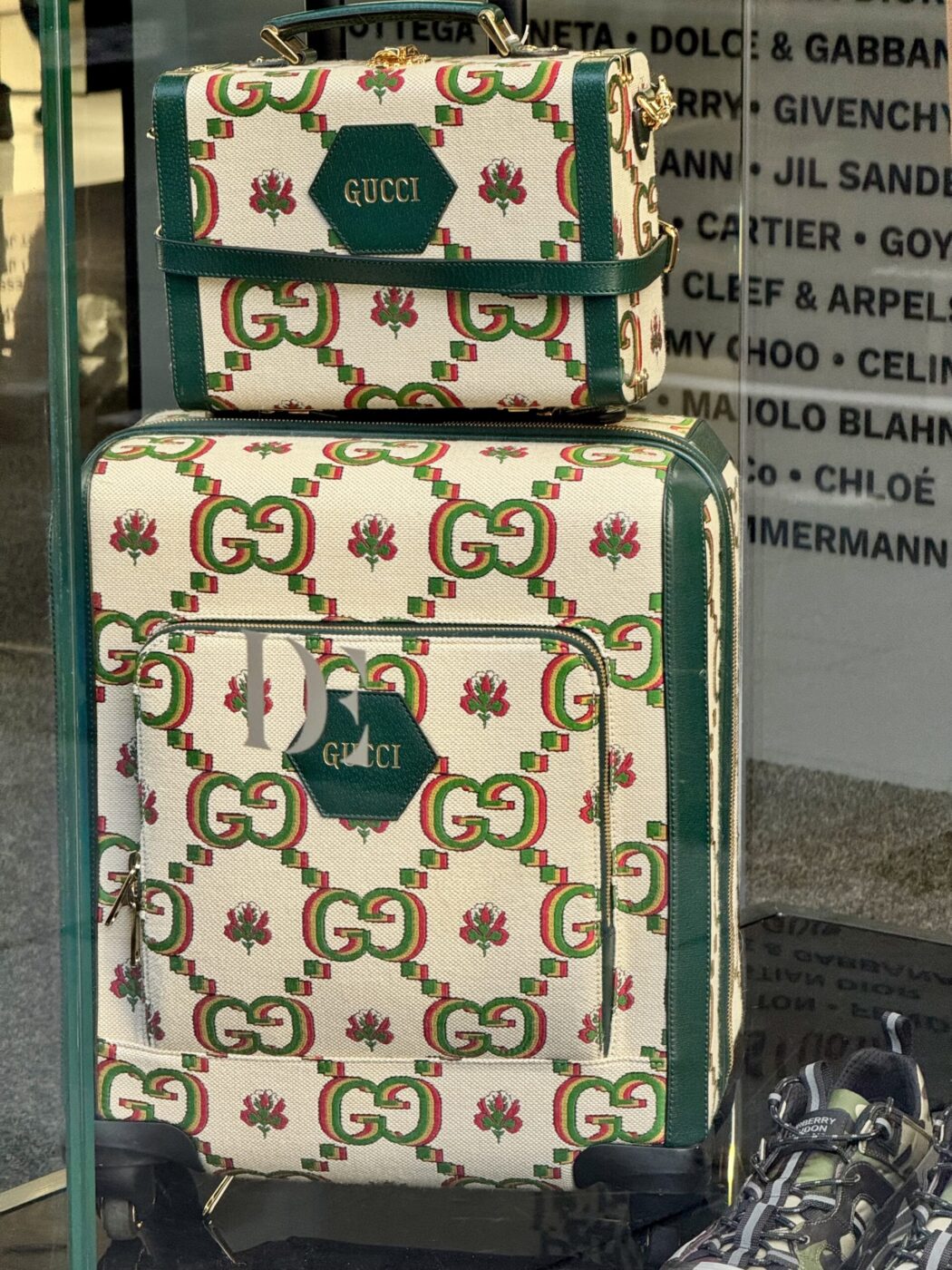 Two Gucci floral-patterned travel bags in a store window; sneakers to the right, luxury brand names visible in the background.
